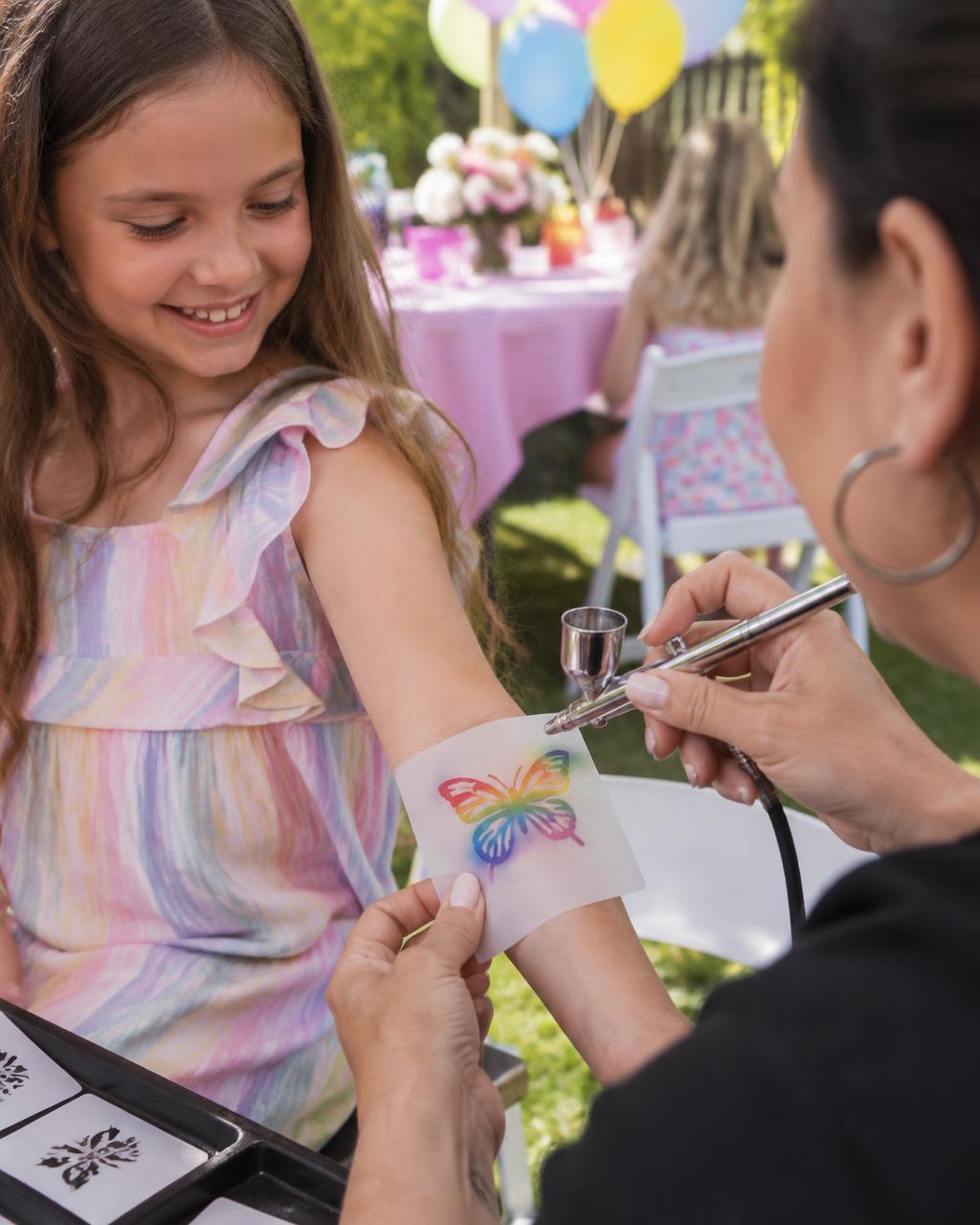 Artist airbrushing a rainbow butterfly temporary tattoo on a girl's arm at a backyard birthday party