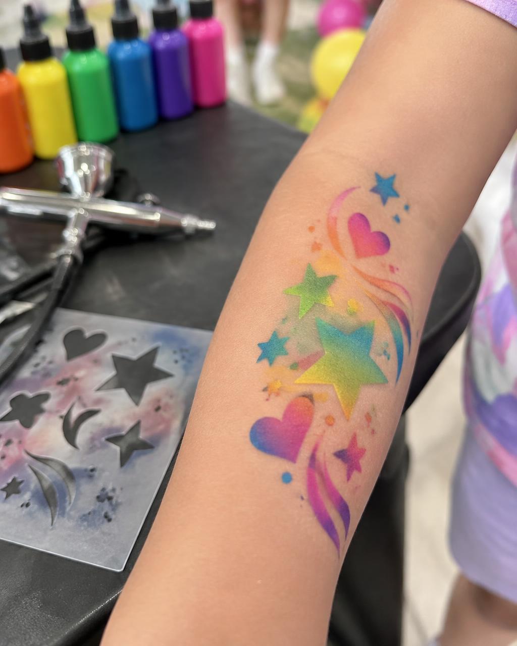 Child's forearm with colorful star and heart temporary airbrush tattoo beside an airbrush gun